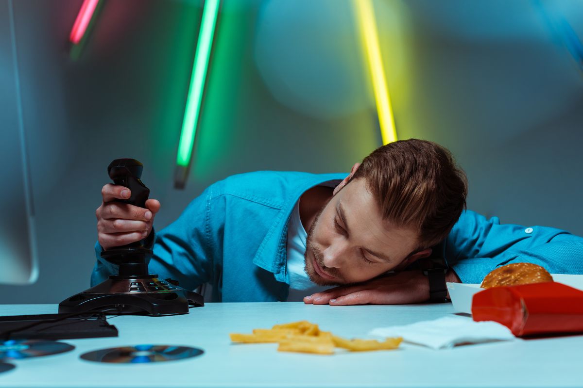 Man in a blue shirt falls asleep at his desk while holding a video game joystick. His head rests on one arm next to scattered fries, a burger, and game discs. The image conveys staying up late gaming and eating fast food until exhaustion.