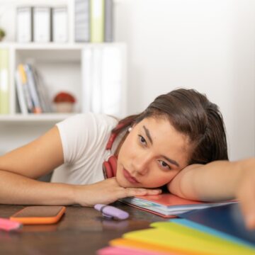 An overwhelmed woman resting her head on a desk, looking drained and unmotivated, with notebooks, a phone, and office supplies in front of her.