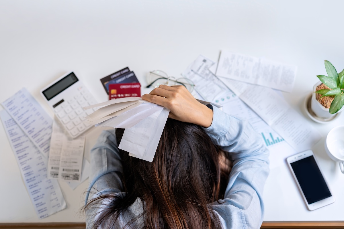 A woman with her head down on a desk surrounded by receipts, credit cards, a calculator, and scattered papers.