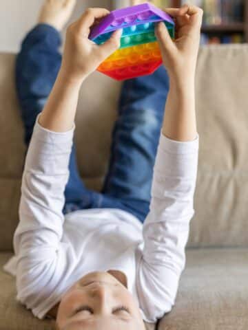 A kid on a sofa with his feet in the air holding a rainbow toy gift.