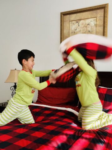 Two kids pillow fighting on a bed in holiday pajamas.